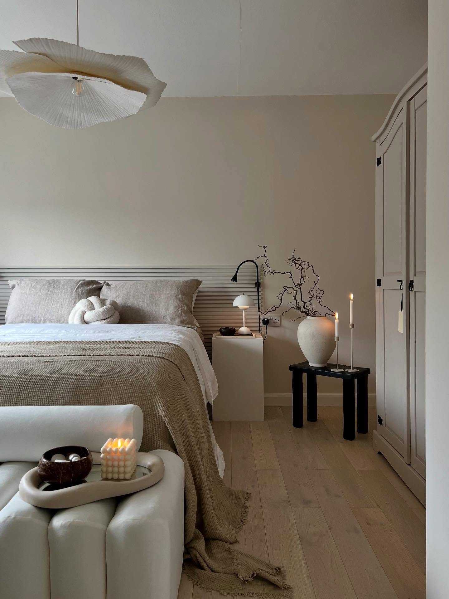 Minimalist bedroom with a slatted neutral headboard, layered beige and white bedding, a modern white wardrobe, and a large sculptural pendant light.