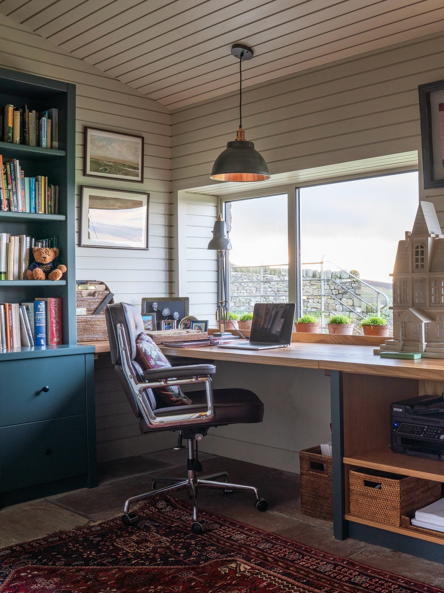 Cozy home office with pale wood plank walls, a large window overlooking a stone wall, a teal bookshelf, and a dark leather office chair over a red patterned rug.