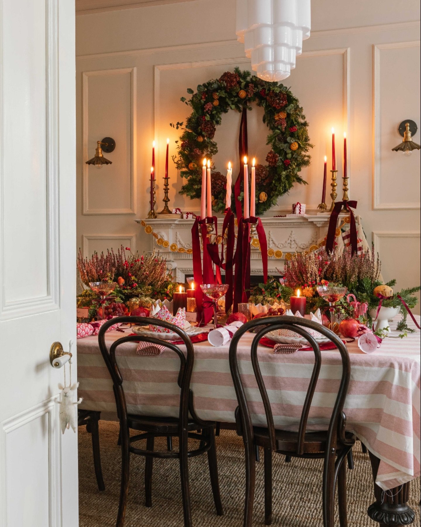 A festive dining room with a table covered in a pink and white striped tablecloth, set with dark plates, heather floral arrangements, and deep red velvet ribbons hanging from the centerpiece candles.