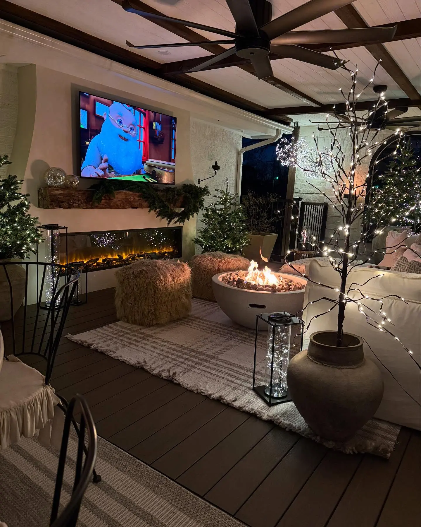 Covered outdoor patio with a wide, linear electric fireplace and a TV mounted above a thick rustic wooden mantel, next to a large circular fire pit.