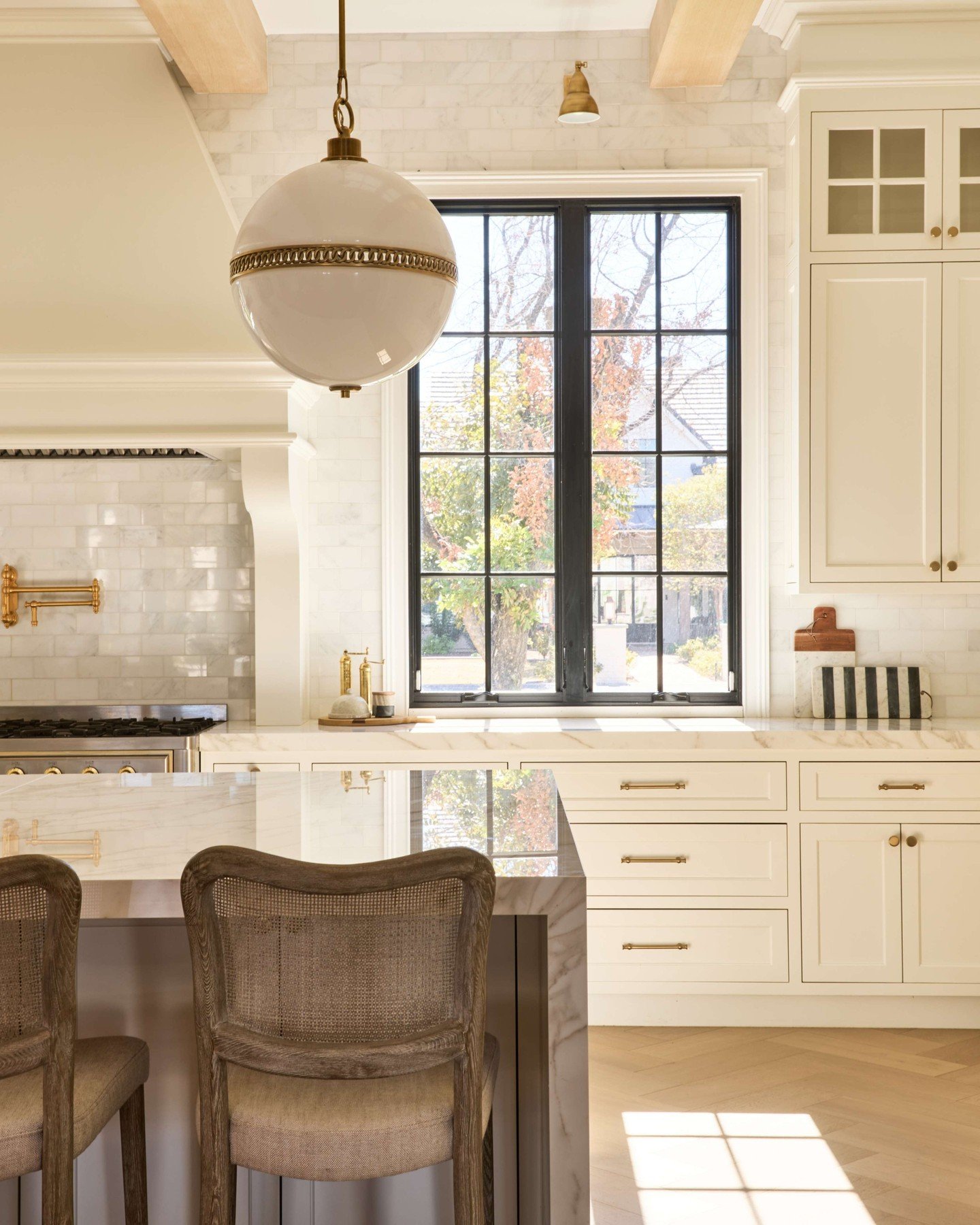 Bright, transitional kitchen with off-white cabinetry and marble counters. A large black-framed grid window is centered on the wall, creating a dramatic contrast with the light marble backsplash.