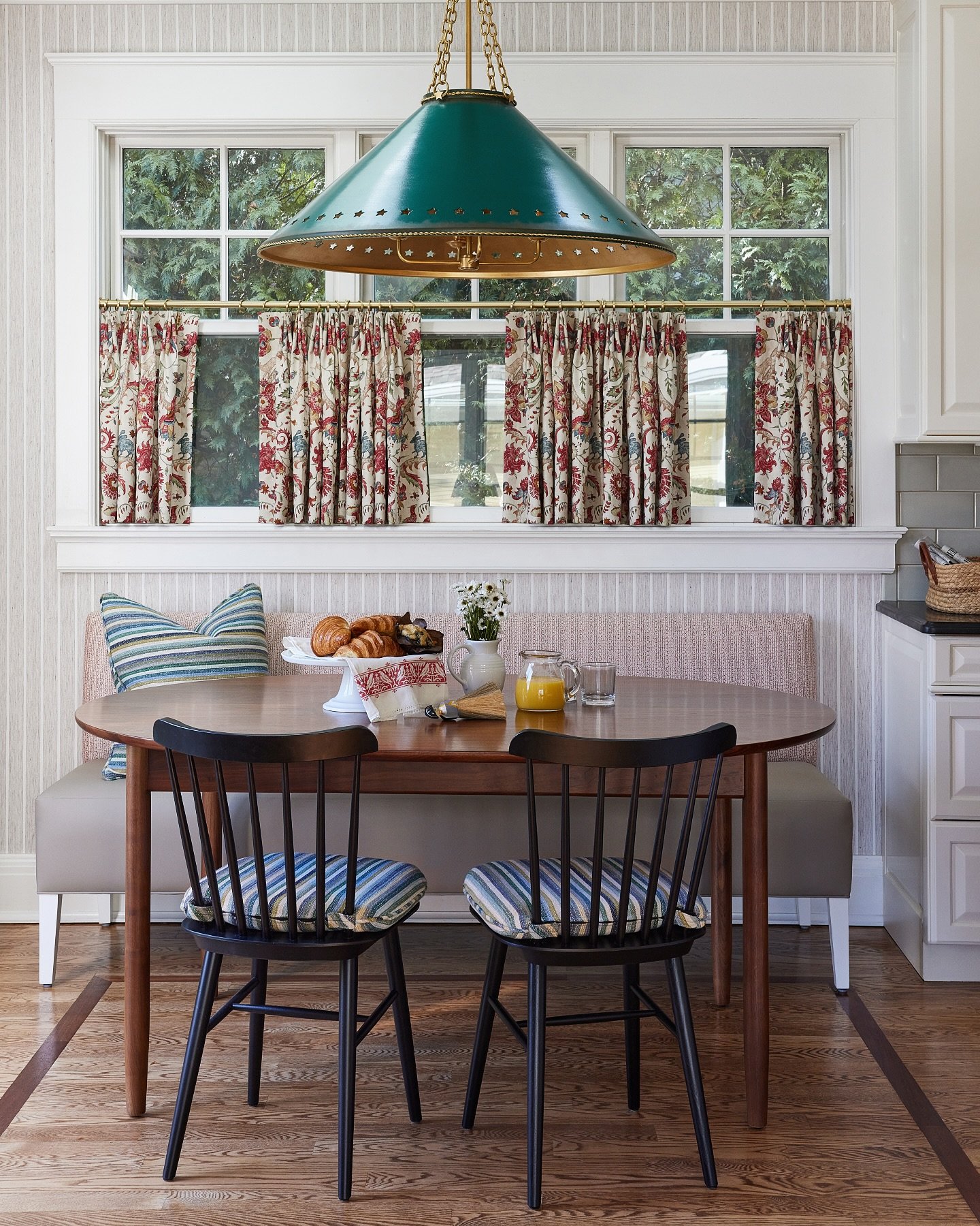 Kitchen breakfast nook with a wooden oval table and two black chairs, featuring traditional red and green floral gathered valance curtains across the window.