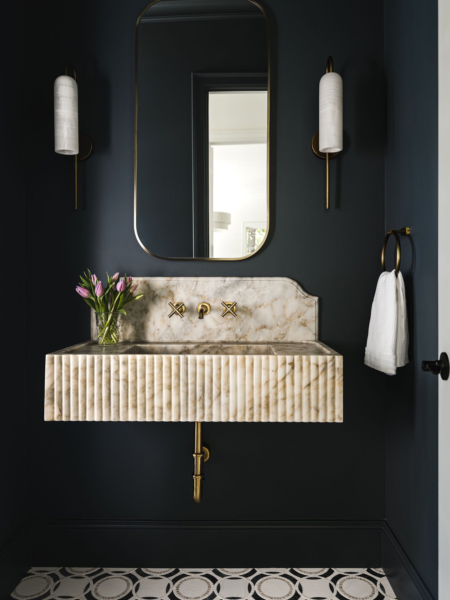 Floating fluted marble sink against a dark blue wall with a gold-framed mirror and sconces. Description: This space immediately draws you in with its bold, dark indigo walls.