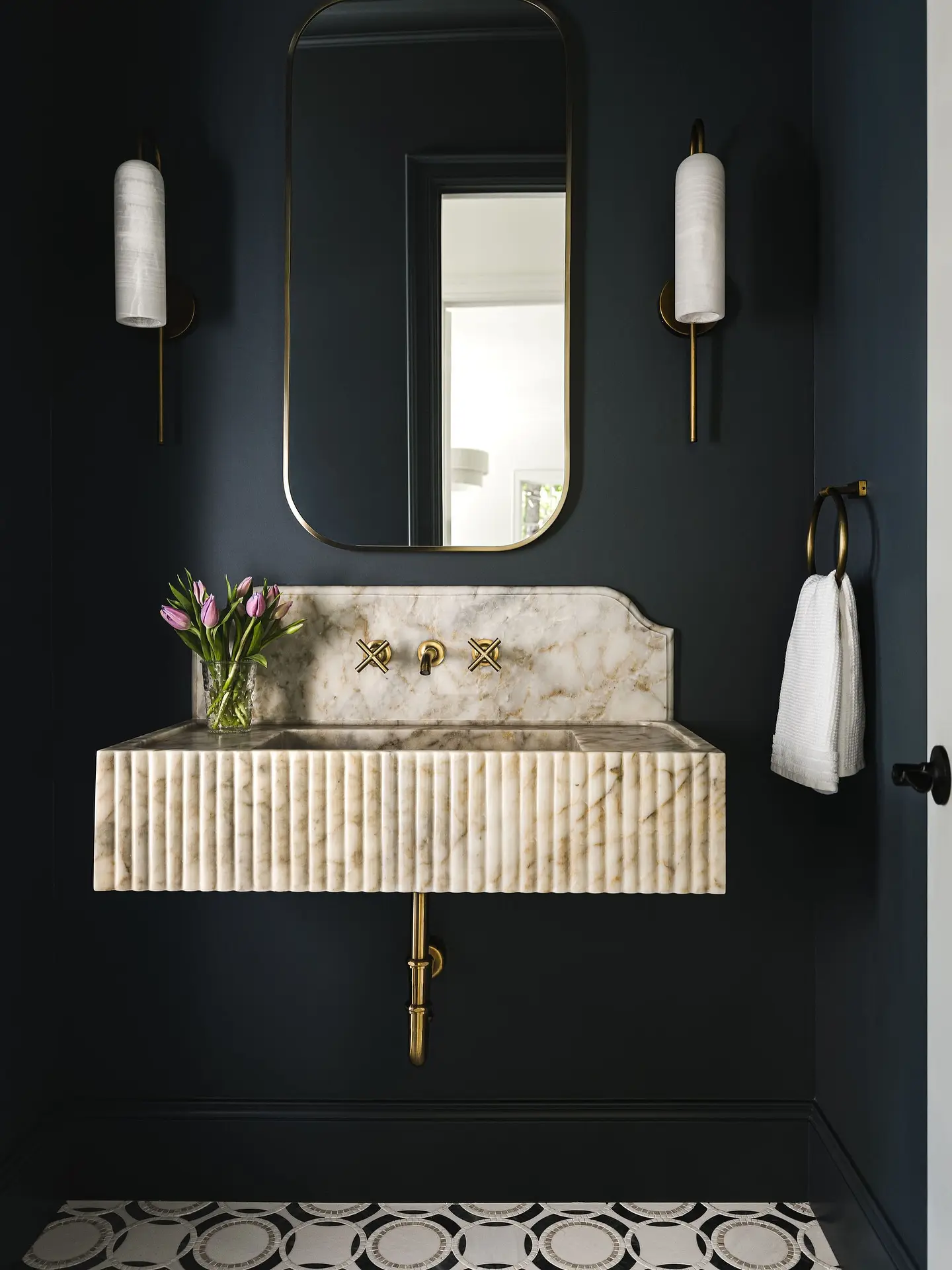 Floating fluted marble sink against a dark blue wall with a gold-framed mirror and sconces. Description: This space immediately draws you in with its bold, dark indigo walls.