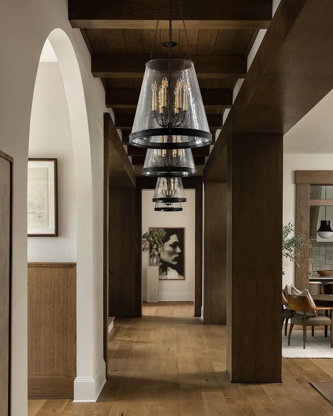 Long, narrow hallway with dark wood ceiling beams, exposed wood columns, light wood flooring, and a series of large seeded glass and black metal pendant chandeliers.