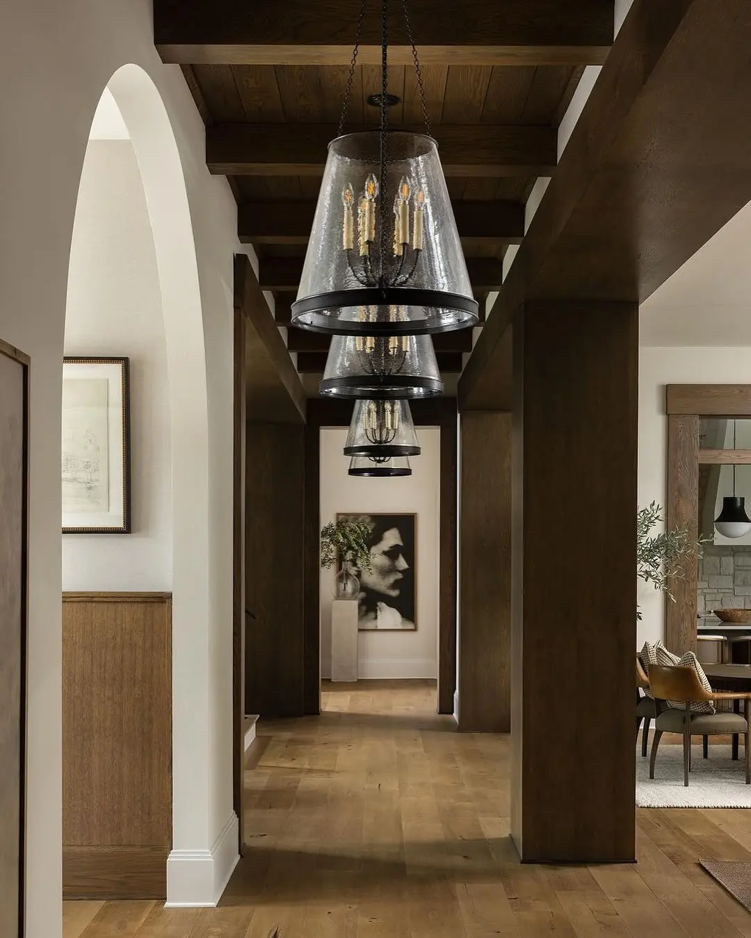 Long, narrow hallway with dark wood ceiling beams, exposed wood columns, light wood flooring, and a series of large seeded glass and black metal pendant chandeliers.