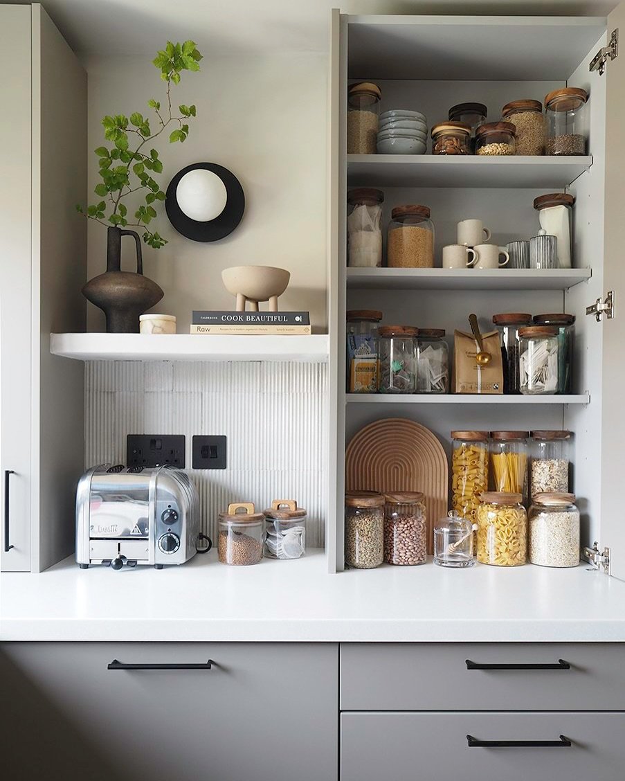 Modern grey kitchen cabinets with an open door revealing organized pantry goods next to a small countertop appliance area.