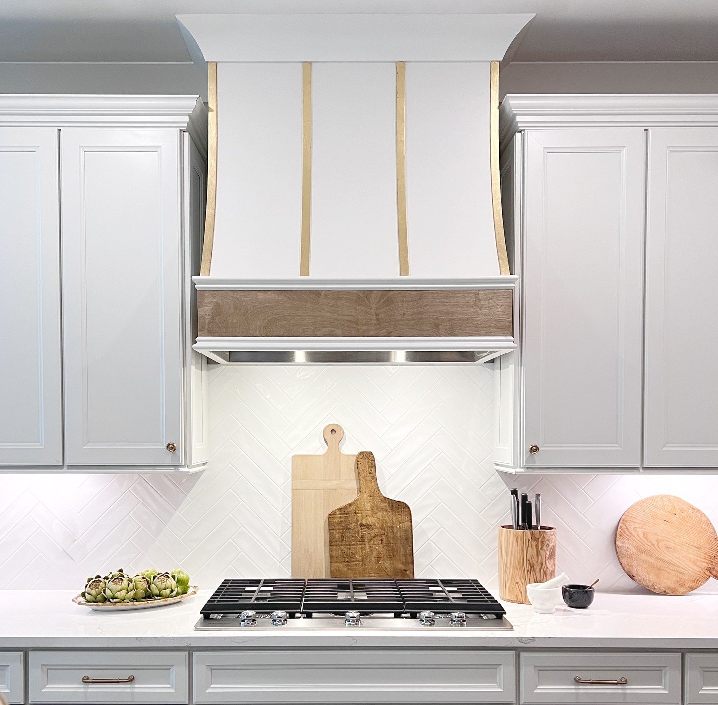 Close-up of a kitchen range area featuring a white chevron subway tile backsplash, a custom white and wood range hood with gold accents, and white quartz countertops.