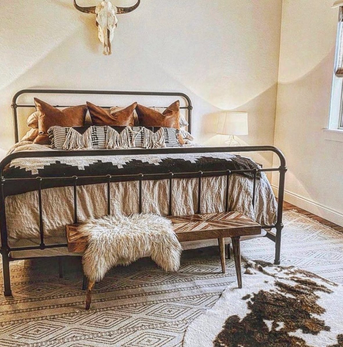A Western bedroom with a black metal bed frame, a patterned rug, leather pillows, and a wooden bench with a sheepskin throw.