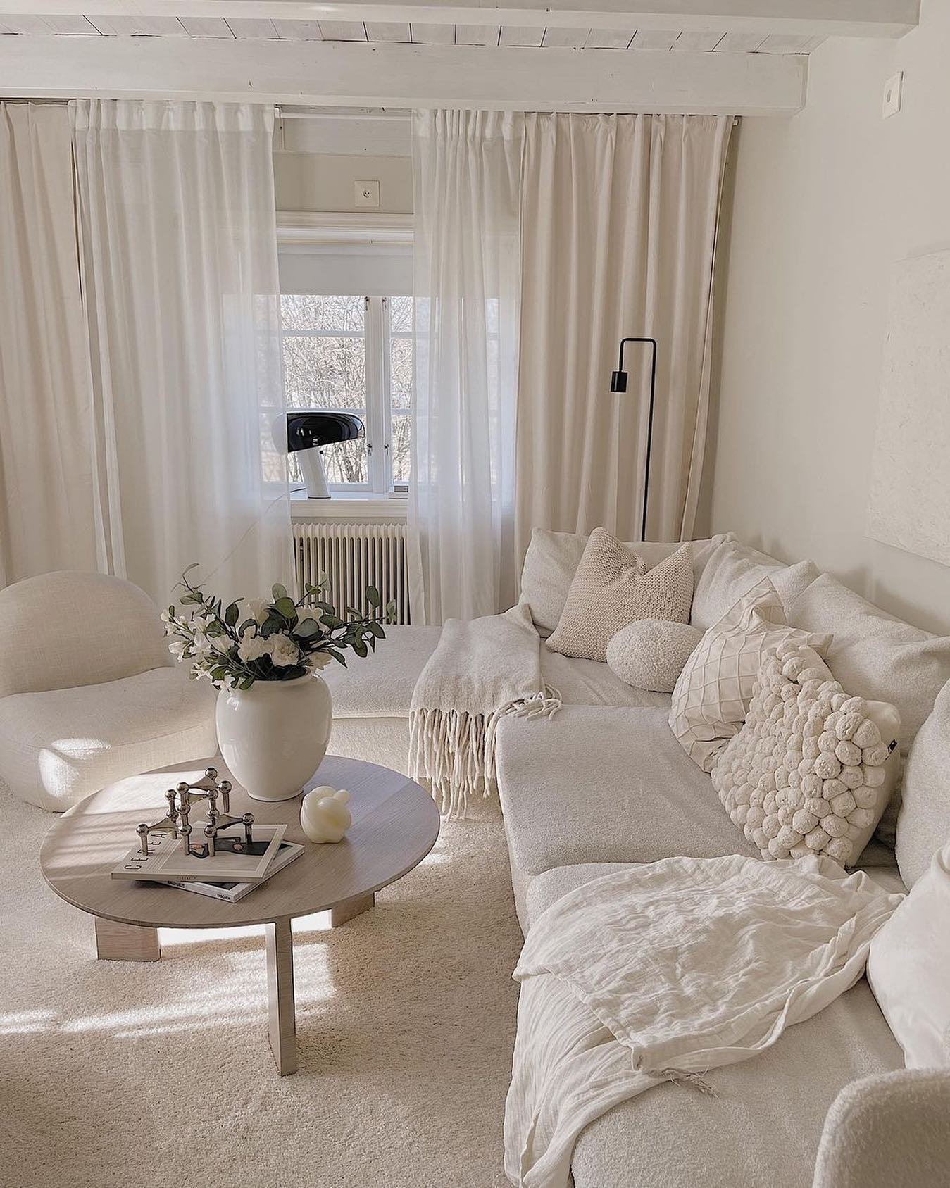 A cozy, light beige living room featuring an oversized sectional, sheer white curtains, a low round coffee table, and a large vase of flowers.