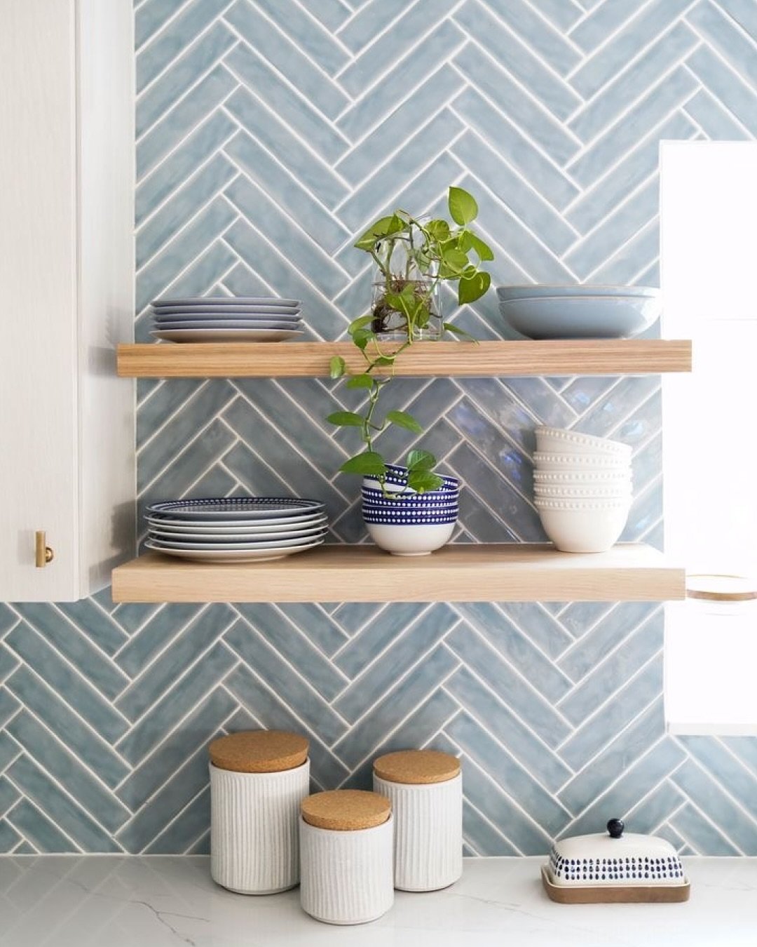 Kitchen backsplash with light blue, elongated subway tiles installed in a crisp herringbone pattern, accented by two floating wooden shelves.