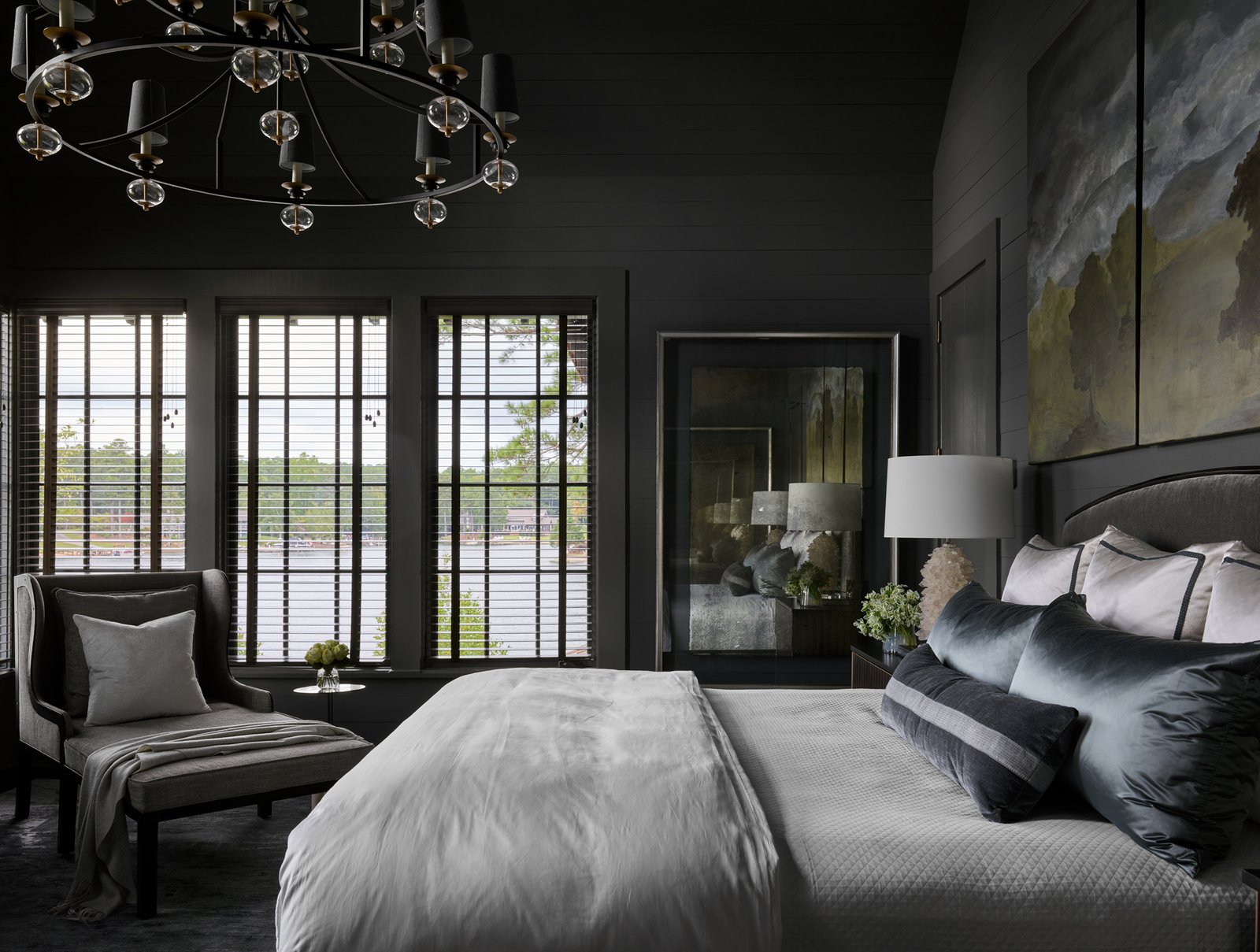 Dark, sophisticated bedroom with charcoal shiplap walls, a wrought iron and crystal chandelier, large windows overlooking a lake, and a grey upholstered bed.