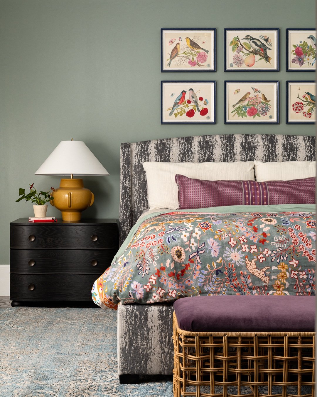 Black wood nightstand with three rounded drawers beside a bed with a floral duvet and colorful bird artwork.