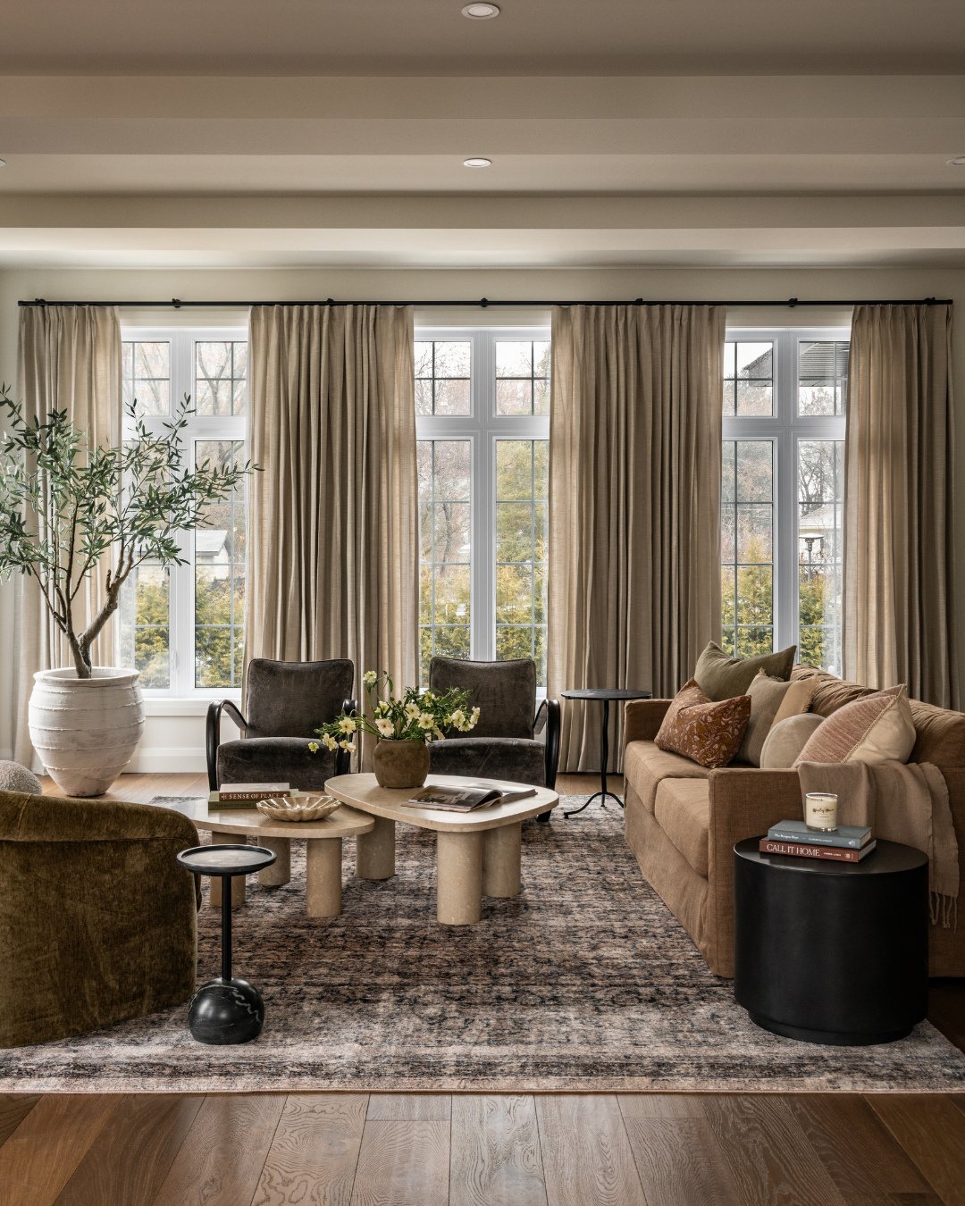 A spacious living room featuring a camel-colored sofa, dark velvet armchairs, a sculptural stone coffee table, and an oversized olive tree by large windows.
