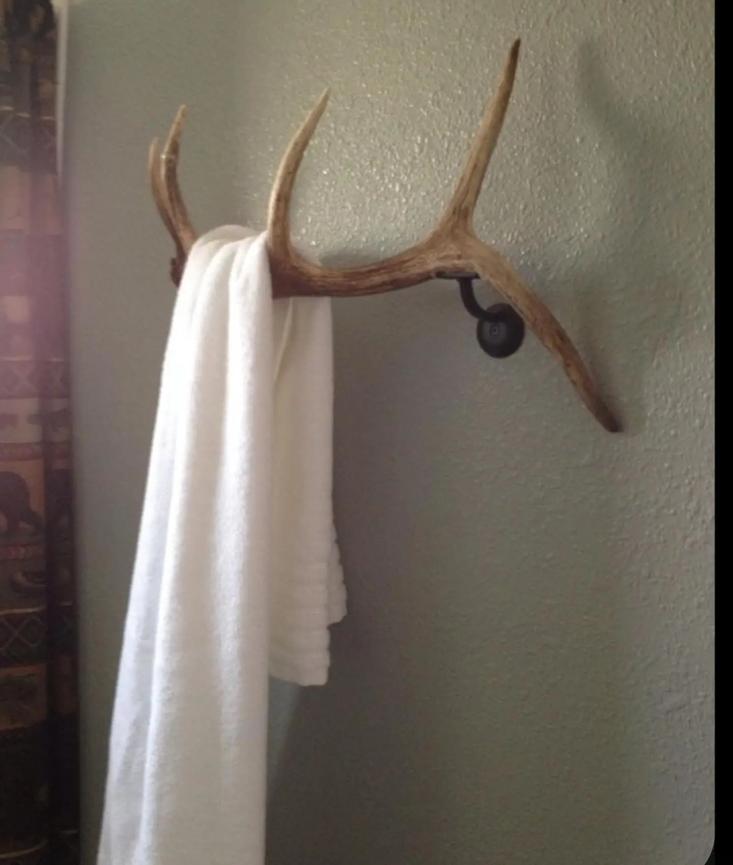 A real or faux deer antler mounted on a pale green textured wall, serving as a unique towel hook and holding a folded white bath towel.