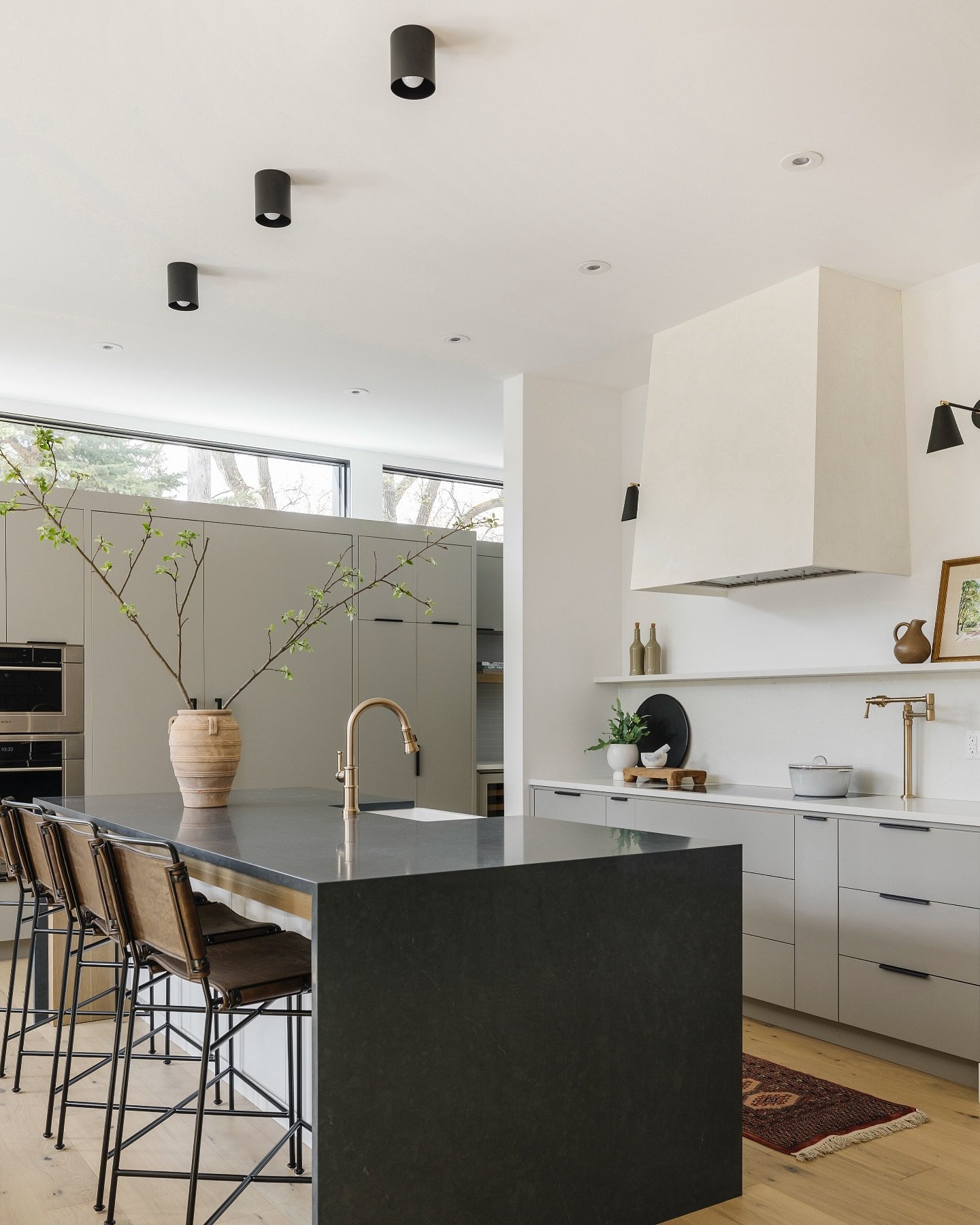 Minimalist kitchen featuring soft matte grey cabinetry, a dramatic dark stone waterfall island, a cream-colored custom range hood, and brass fixtures.