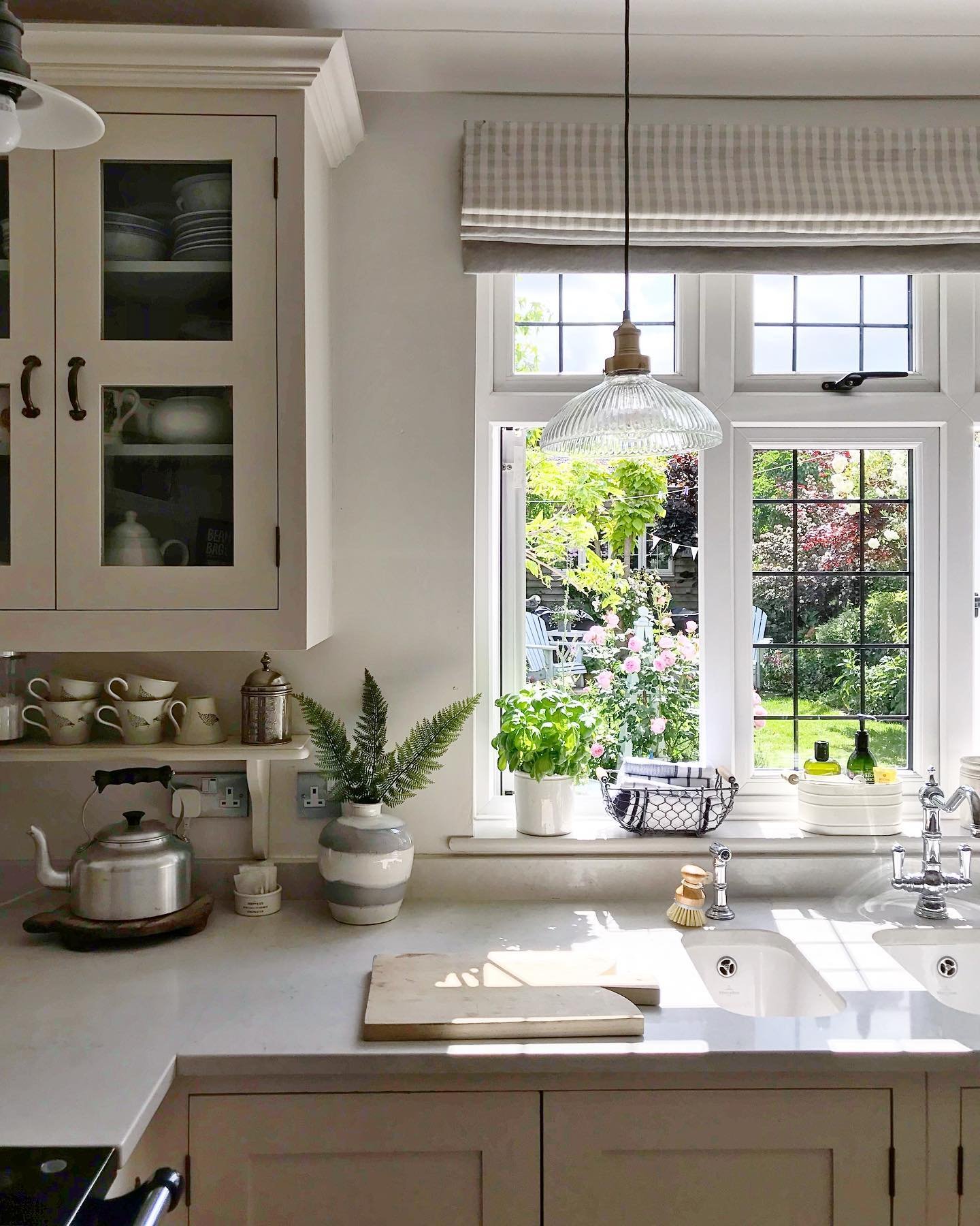 Cozy kitchen with pale grey cabinets and a double white sink. A white-framed casement window is open, offering a lush view of a blooming garden. A gingham Roman shade is partially drawn above.