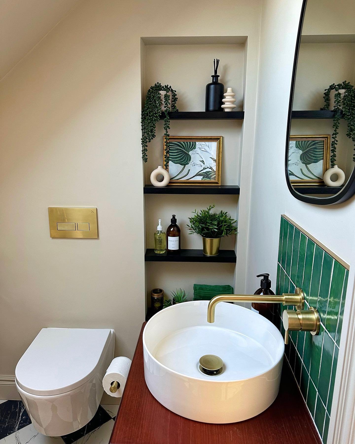 A small recessed wall niche with three dark black floating shelves, styled with a framed print, potted plant, and decorative ceramic vases next to a bathroom sink.