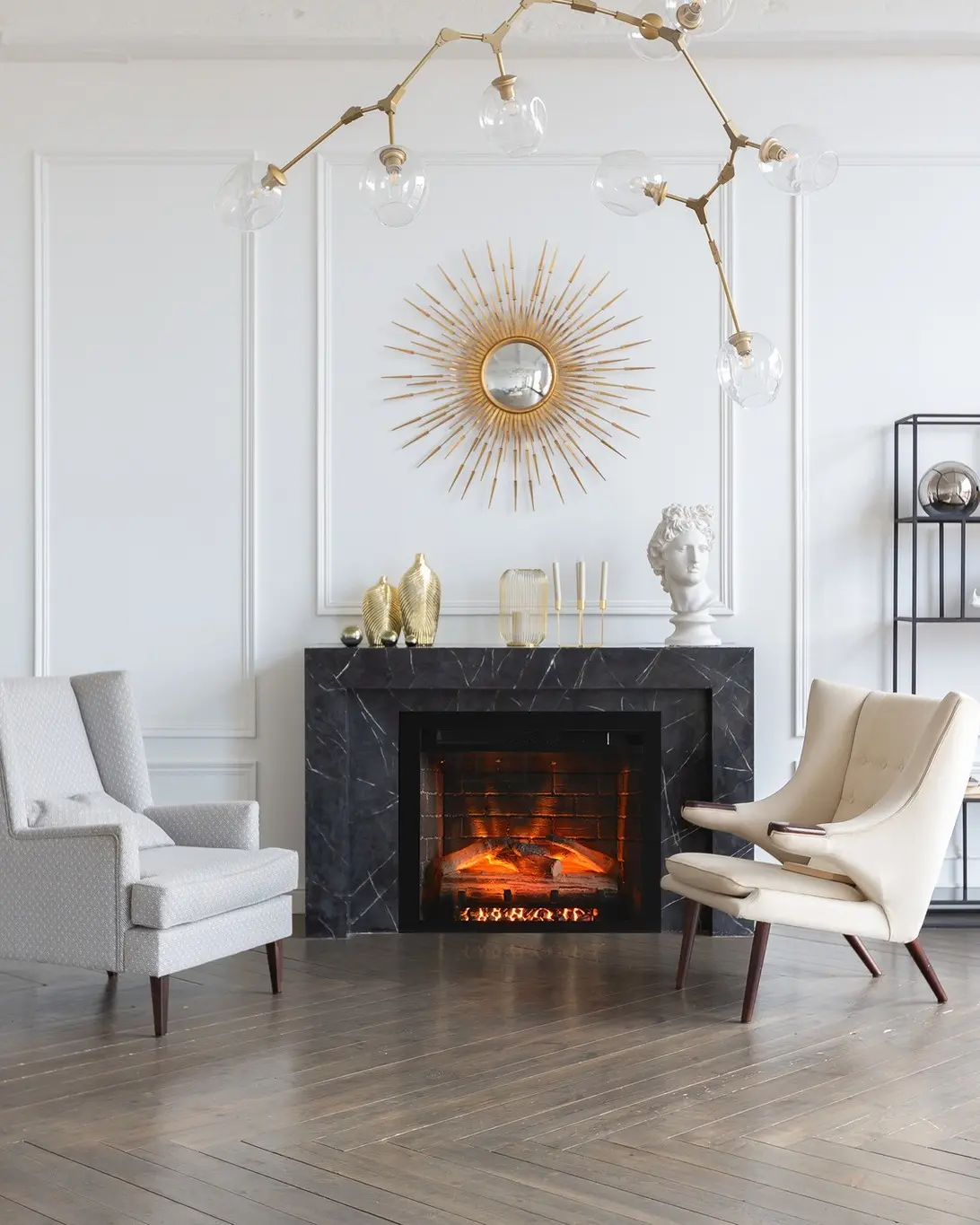 Traditional electric fireplace with a black marble surround and hearth, featuring a warm flame effect, centered on a white wall with wainscoting and a large gold sunburst mirror.