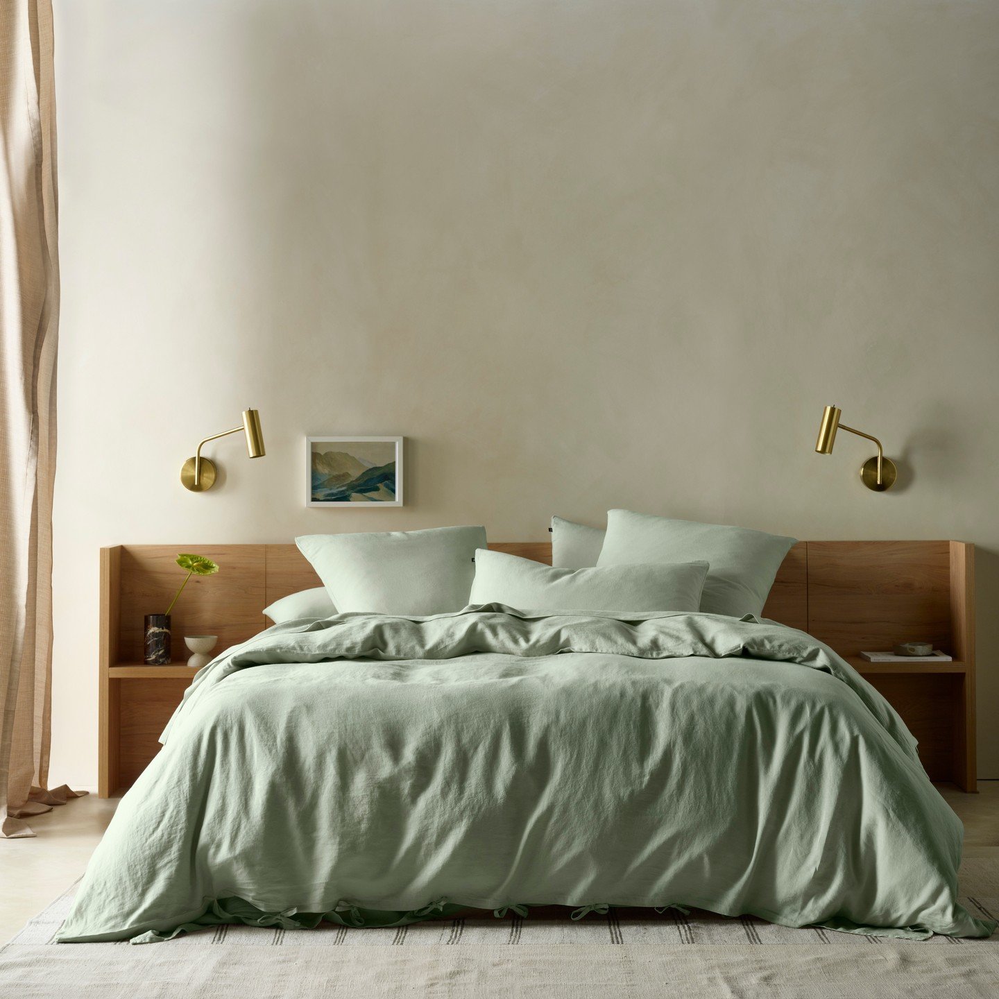 Minimalist bedroom with light textured walls, a wooden headboard, brass sconces, and soft sage green linen bedding.