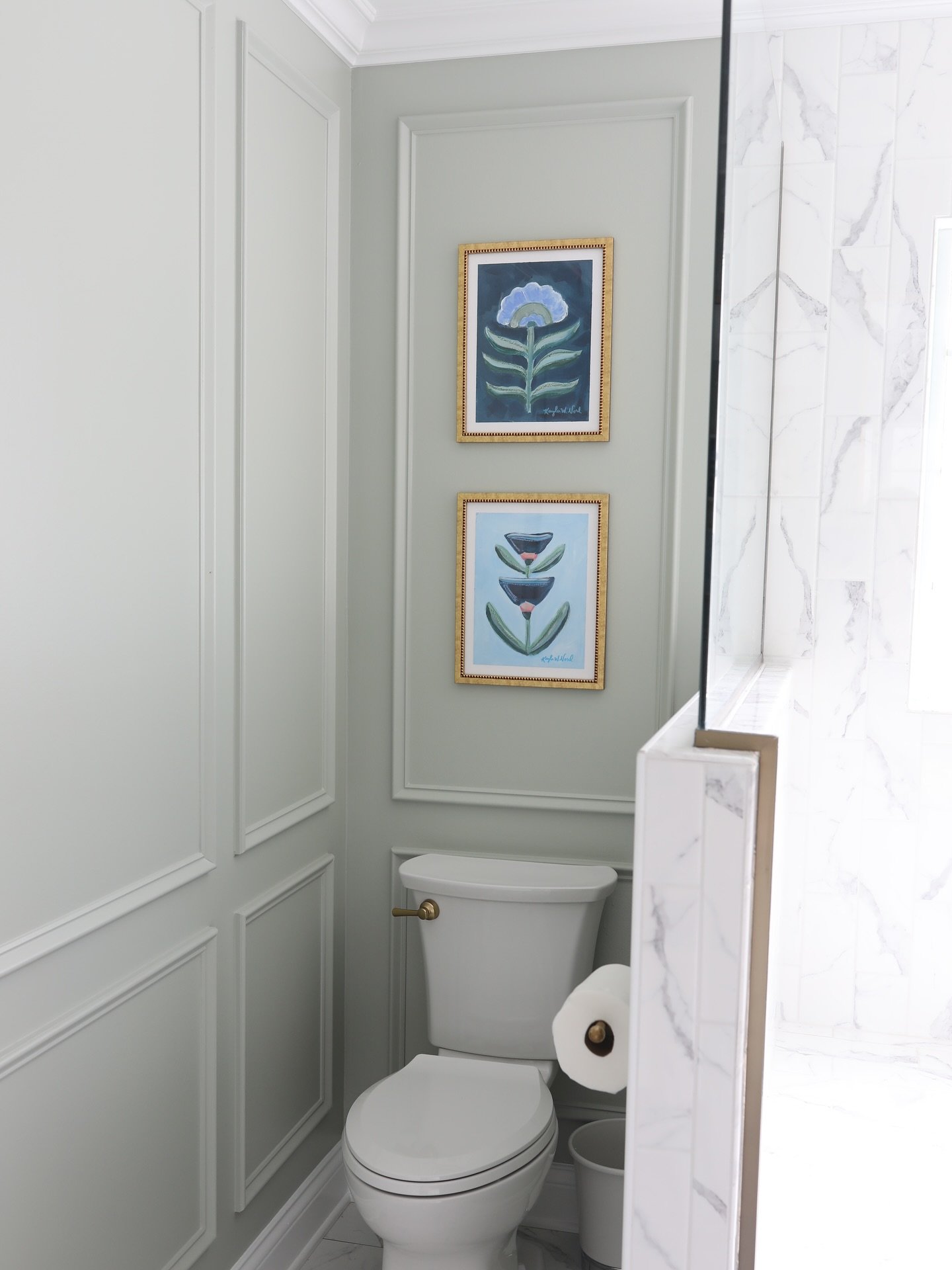Two vertically stacked, gold-framed abstract floral prints over a toilet, set within a light sage-green wall with surrounding white paneling.