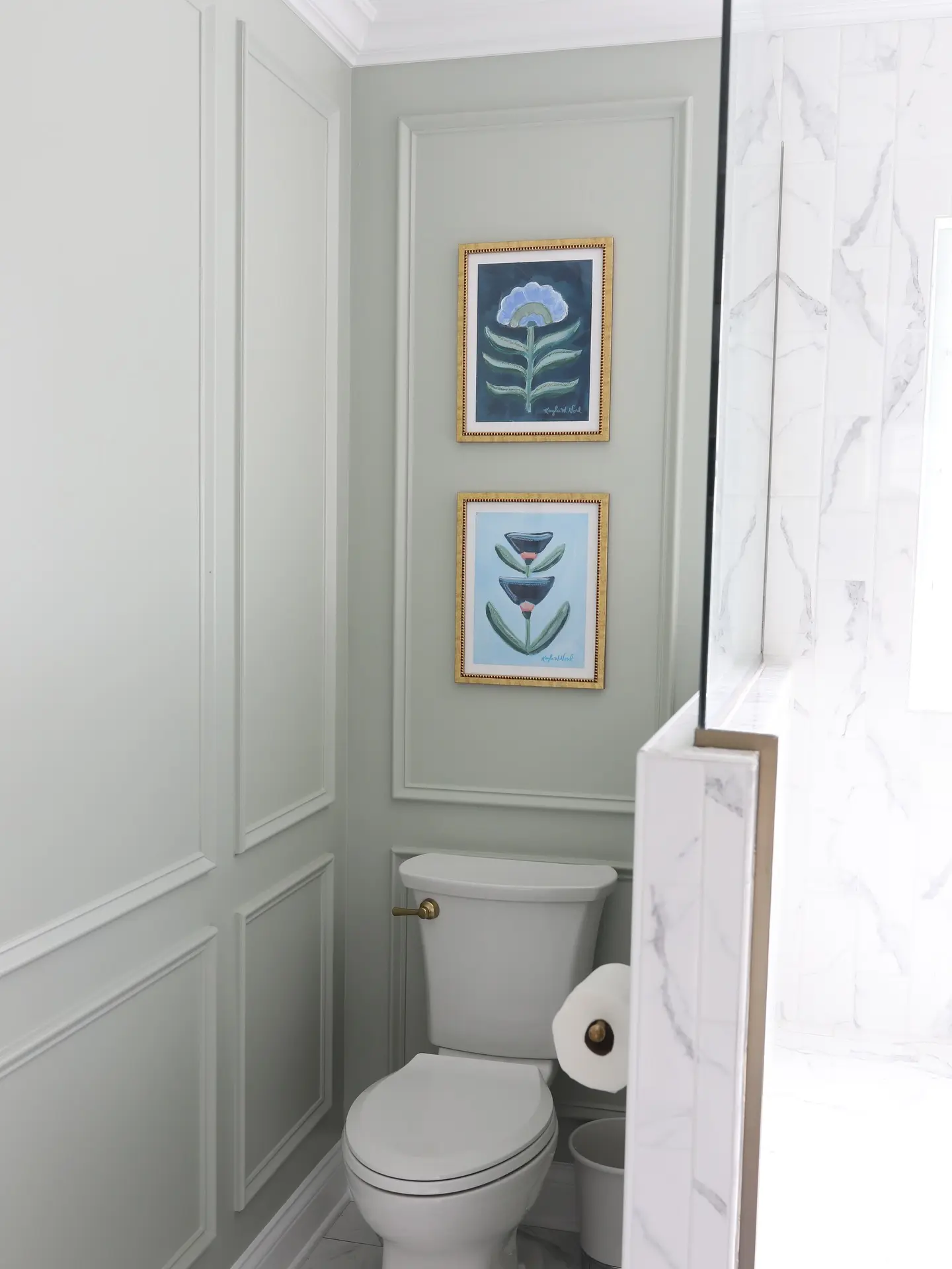 Two vertically stacked, gold-framed abstract floral prints over a toilet, set within a light sage-green wall with surrounding white paneling.