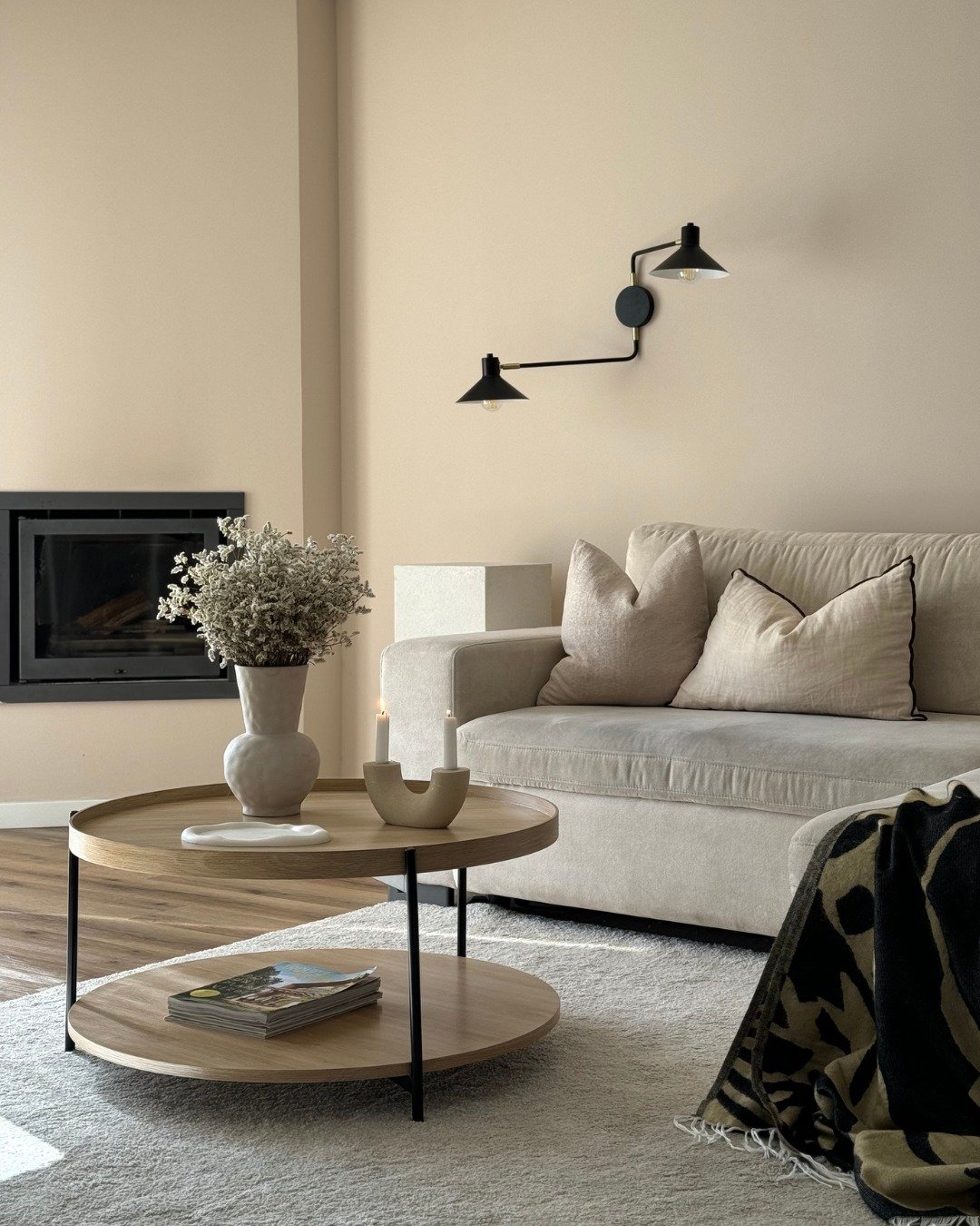 A close-up of a modern beige living room featuring a light linen sofa, a two-tiered wooden coffee table, a black fireplace, and a black articulated wall sconce.