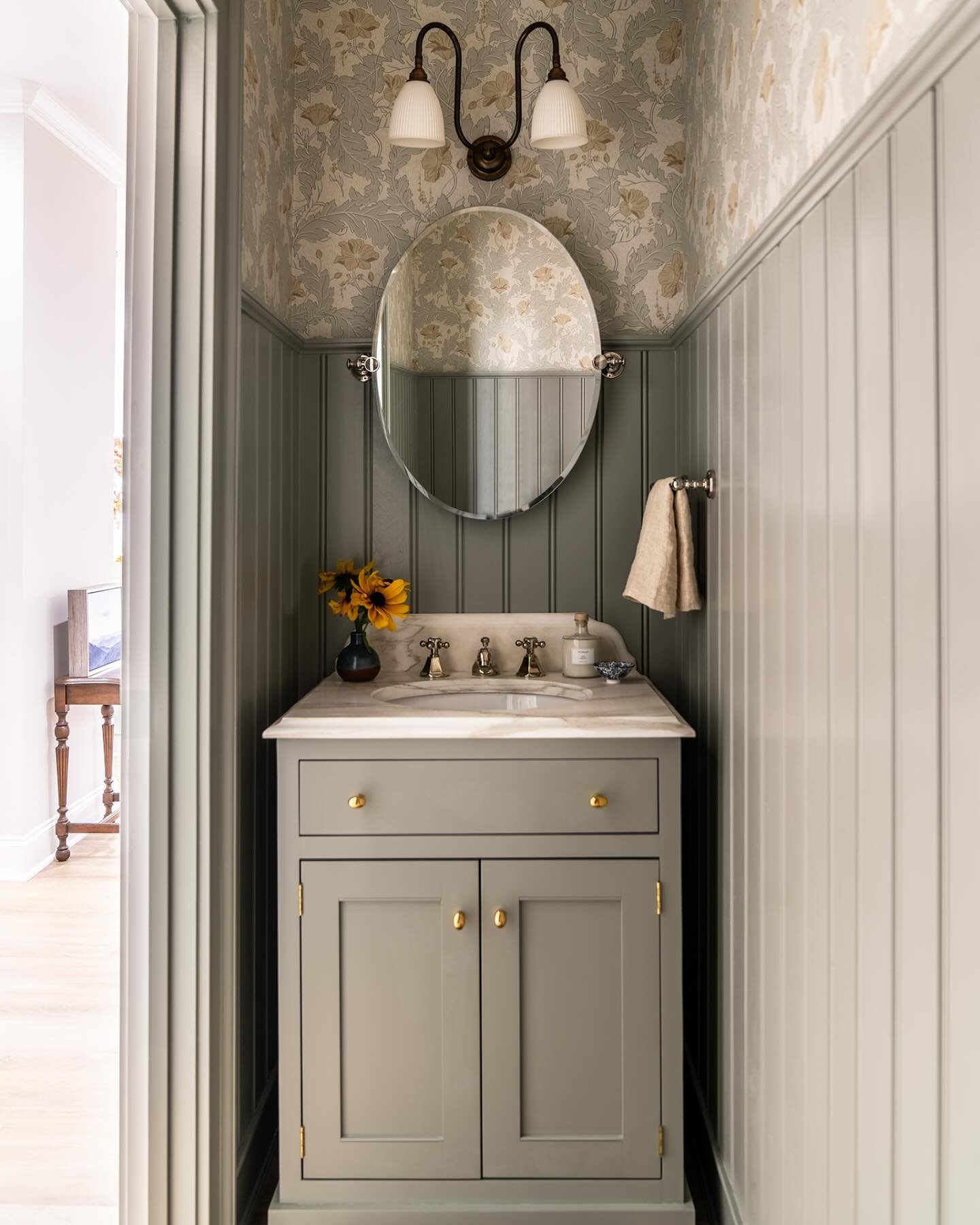 Narrow powder room with sage green beadboard paneling and a simple floral wallpaper above a marble vanity.
