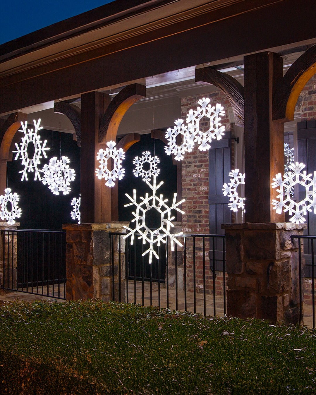 Large, bright white LED snowflake motifs hang at varying heights from the wooden beams of a covered porch with stone columns and a black railing.