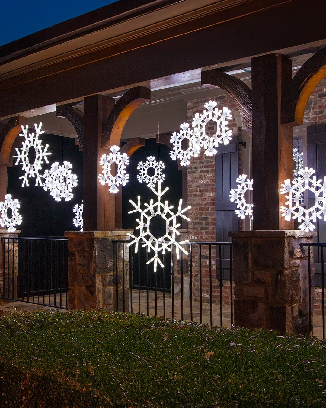 Large, bright white LED snowflake motifs hang at varying heights from the wooden beams of a covered porch with stone columns and a black railing.