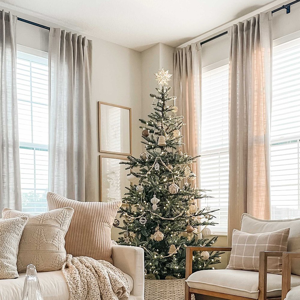 Sheer, floor-length neutral linen blend curtains hung with black rod hardware in a light-filled living room with a cream sofa and Christmas tree.