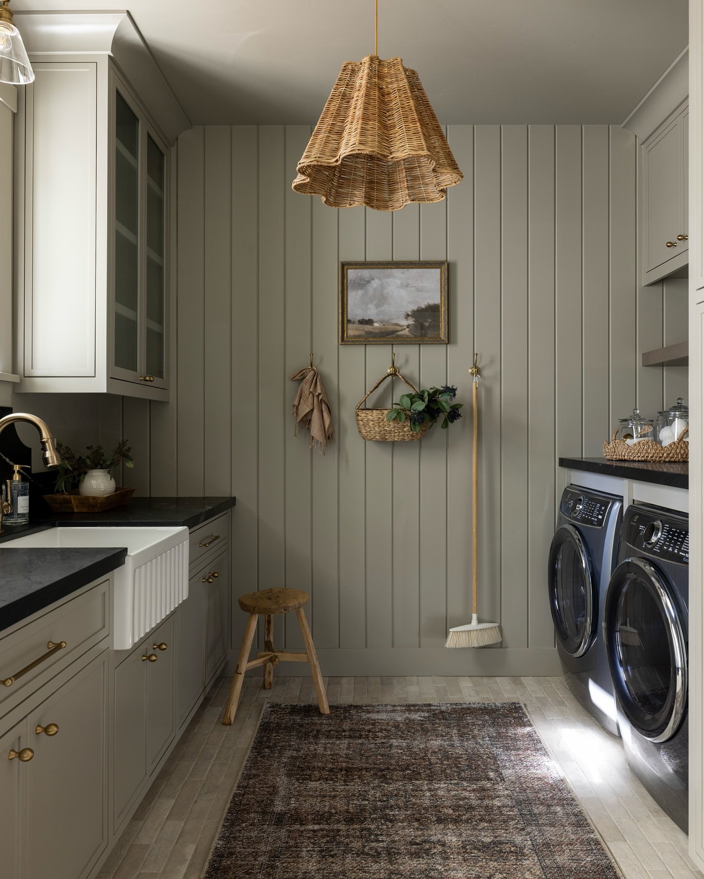 A narrow galley-style laundry room painted in a warm sage-green/greige with vertical shiplap paneling. It features a farmhouse sink, dark countertops, blue washer and dryer, and a woven pendant light.