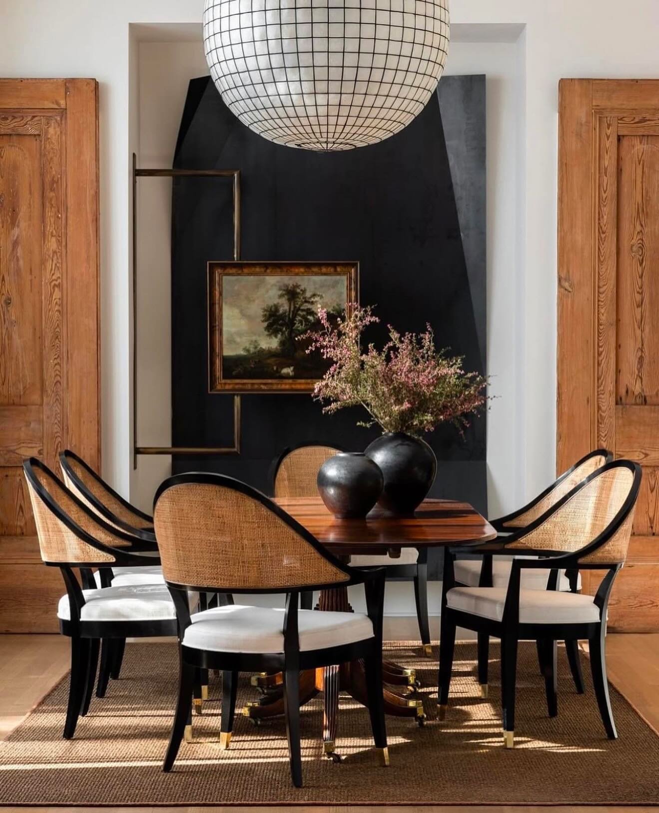 A chic dining area featuring a round pedestal table, black and rattan dining chairs, set against a black and white geometric wall panel flanked by tall wooden doors.