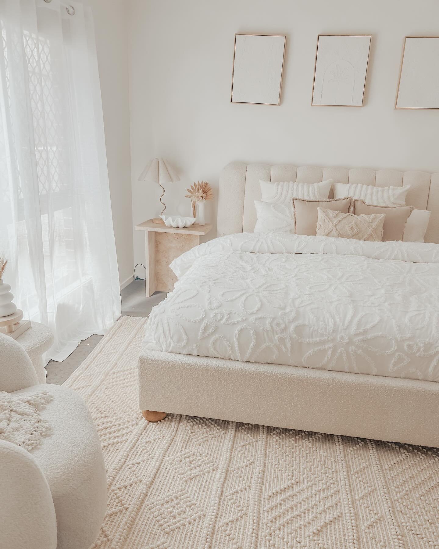 All-white and cream bedroom featuring a textured bouclé bed frame and chair, a highly textured geometric rug, and three white framed art pieces above the bed.