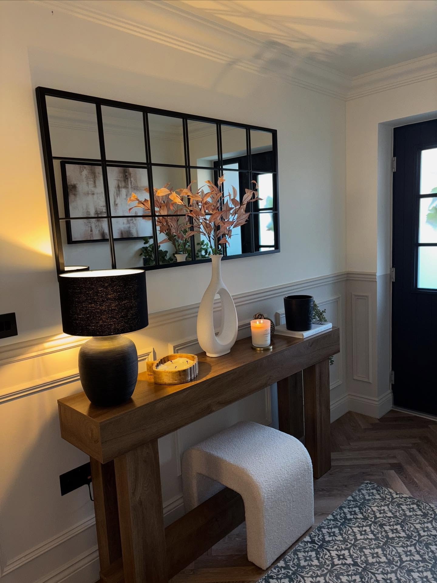 A wooden, waterfall-style console table with a black lampshade, a white vase, and a large black windowpane mirror above it.