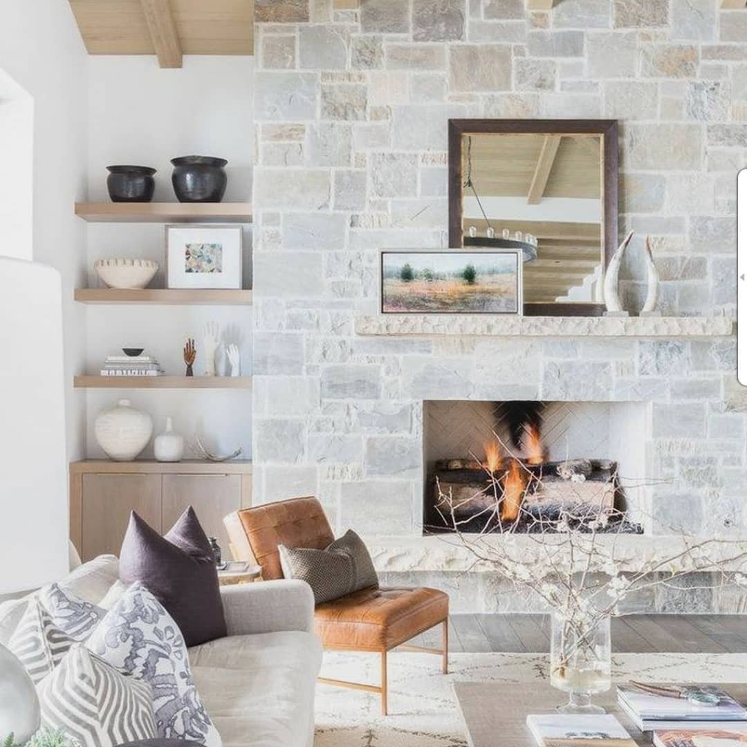 Living room with a full wall of light gray stacked stone, floating wood shelves, a leather armchair, and a wood-framed mirror above the mantel.