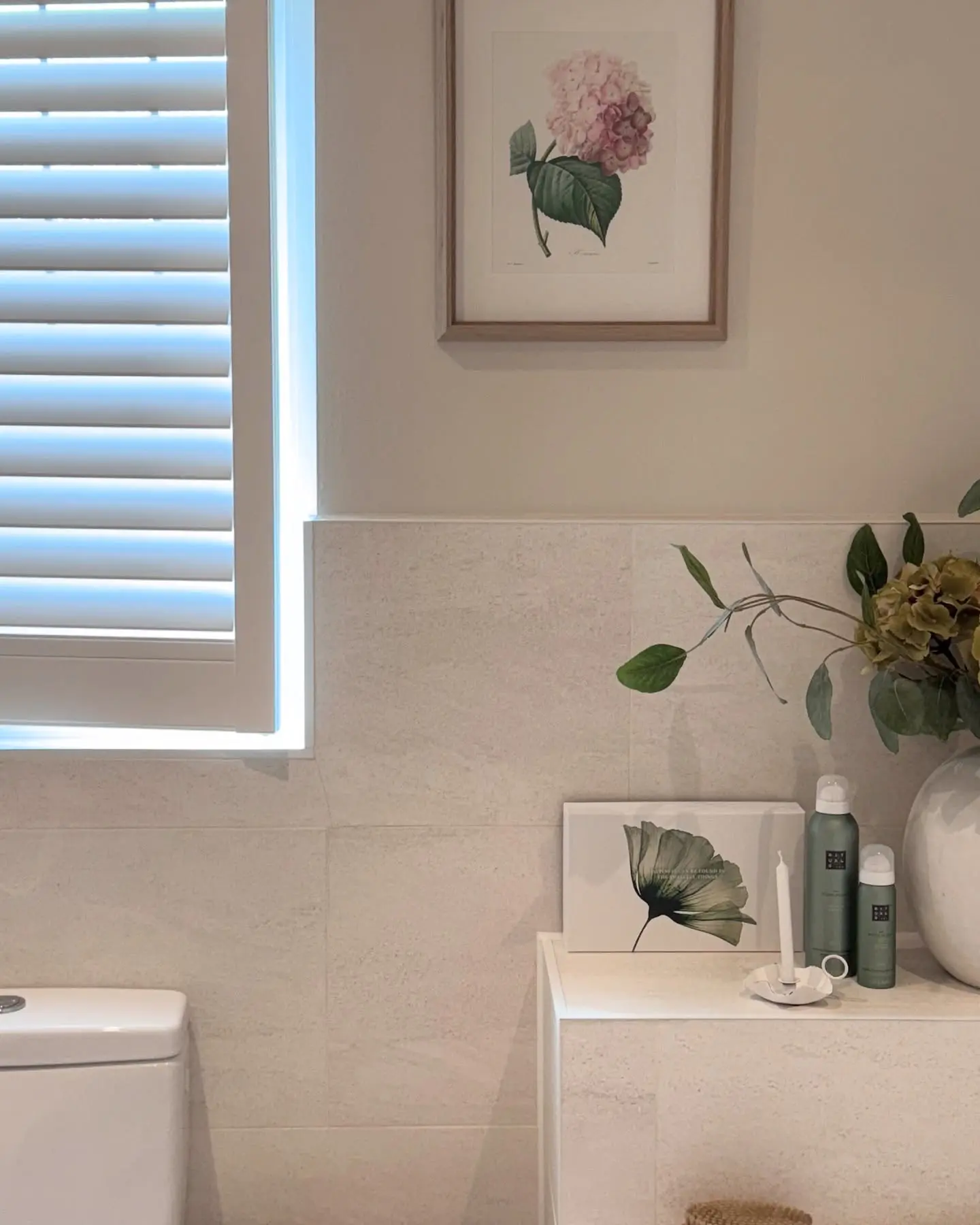 Close-up of a bright, neutral bathroom with light tile, featuring a framed pink hydrangea print and a small black and white floral canvas.