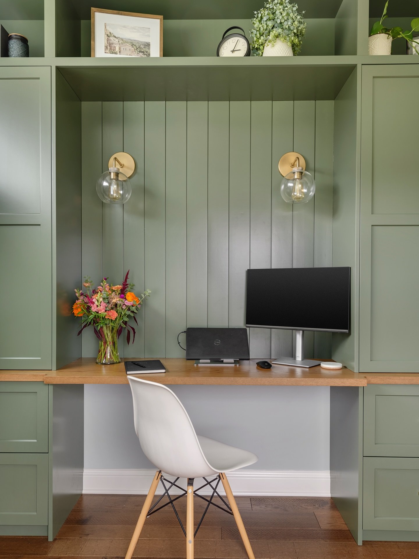 Home office built-in featuring sage green cabinetry, a light wooden desk surface, two brass and glass globe sconces, and a modern white chair.