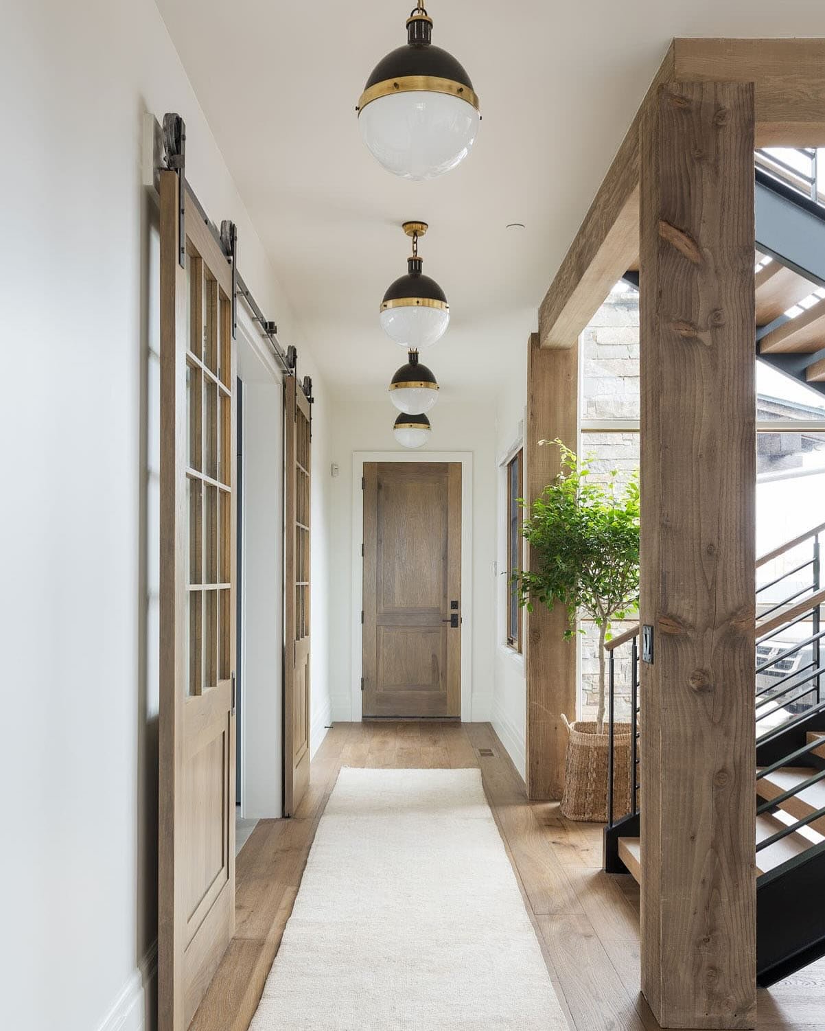 Light and airy hallway with wood floors, natural wood barn doors, a cream runner rug, and a row of black and brass globe pendant lights.