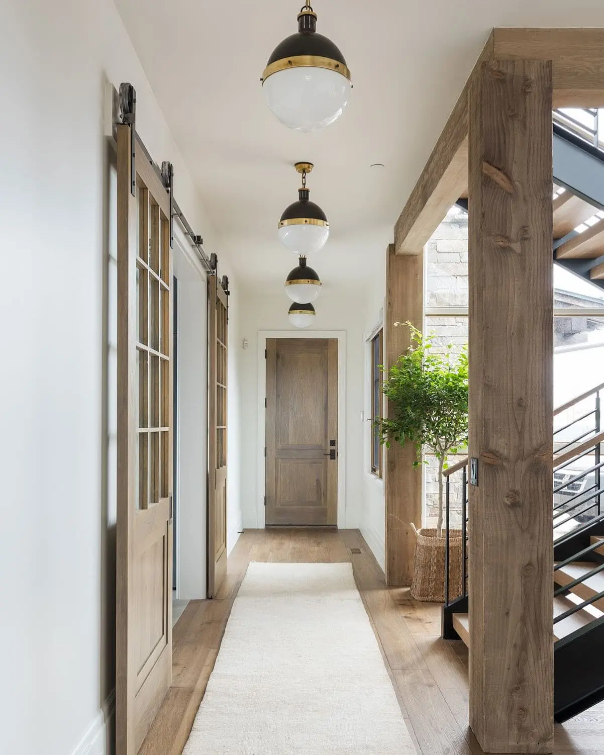 Light and airy hallway with wood floors, natural wood barn doors, a cream runner rug, and a row of black and brass globe pendant lights.
