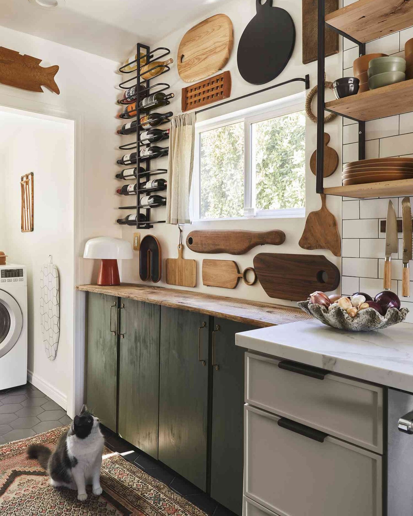 Eclectic kitchen with dark green cabinets, a vertical metal wine rack, and various wooden cutting boards hung as wall art.