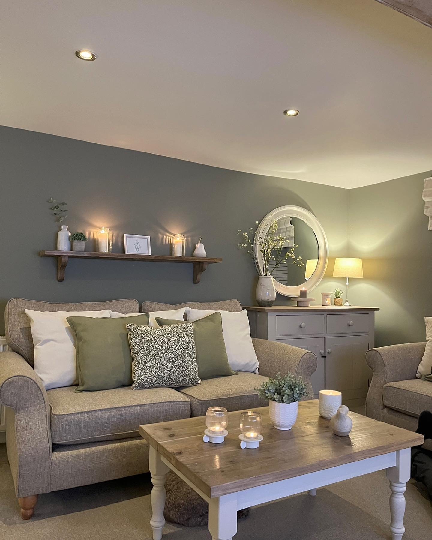A small, cozy living room illuminated by soft lamplight, featuring a taupe sofa against a dark sage-green accent wall.