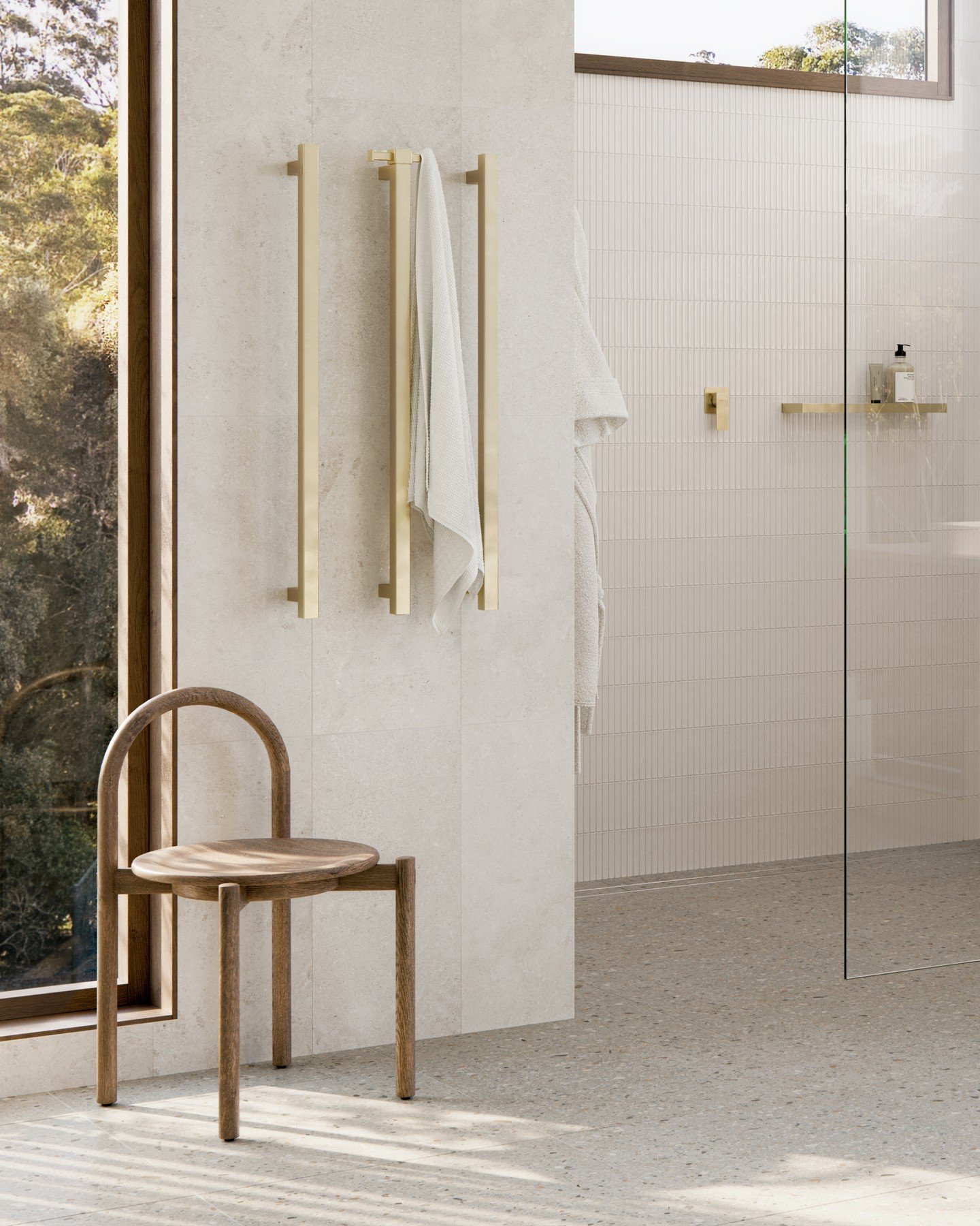 Two sleek, vertical, brushed brass heated towel rails installed on a cream-colored stone tile wall in a light-filled bathroom.