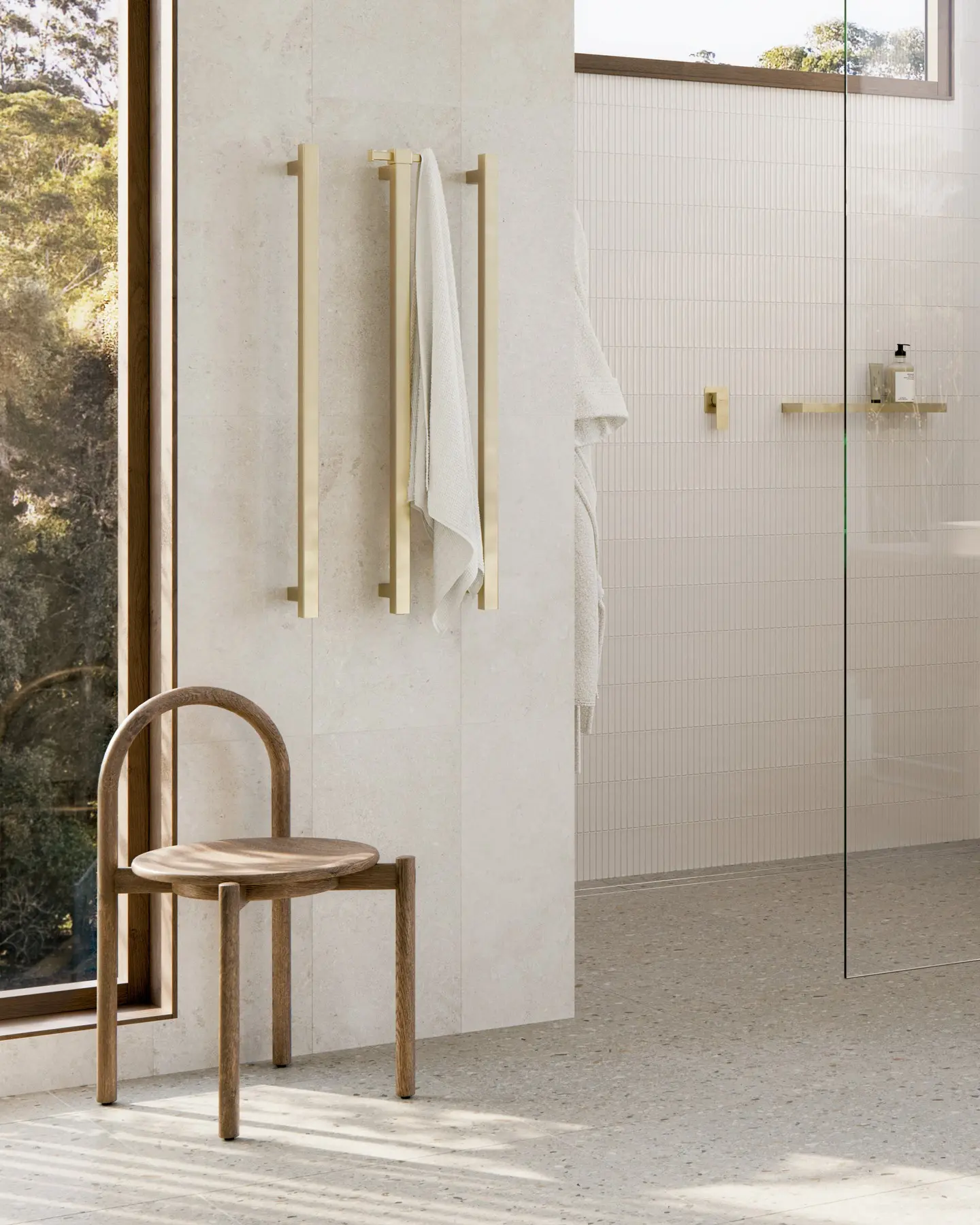 Two sleek, vertical, brushed brass heated towel rails installed on a cream-colored stone tile wall in a light-filled bathroom.