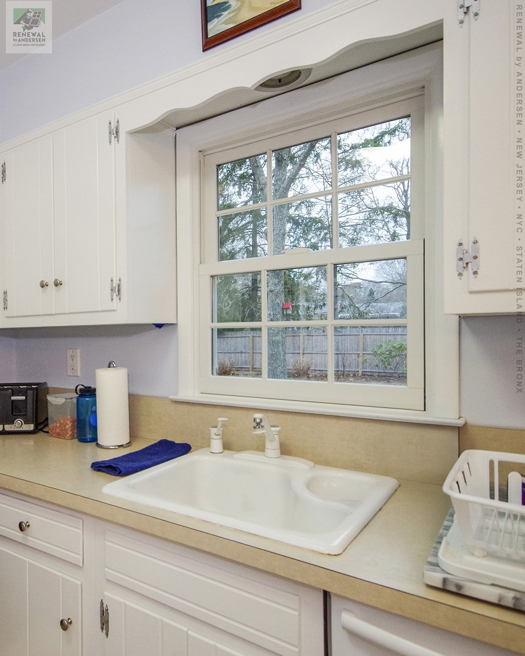 Traditional kitchen setup featuring white shaker cabinets, a light beige countertop, and a white framed grid window centered above a large porcelain sink.