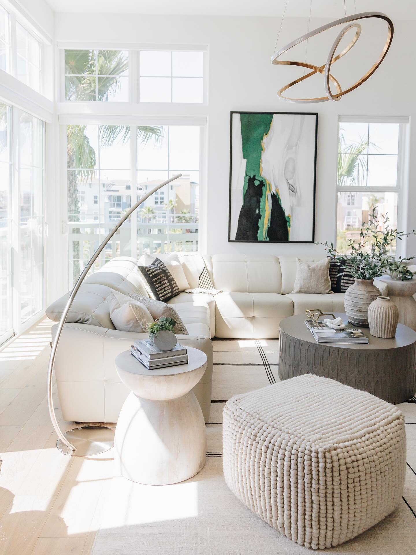 Sunlit living room with white leather sectional, round stone coffee tables, and abstract green and black artwork.