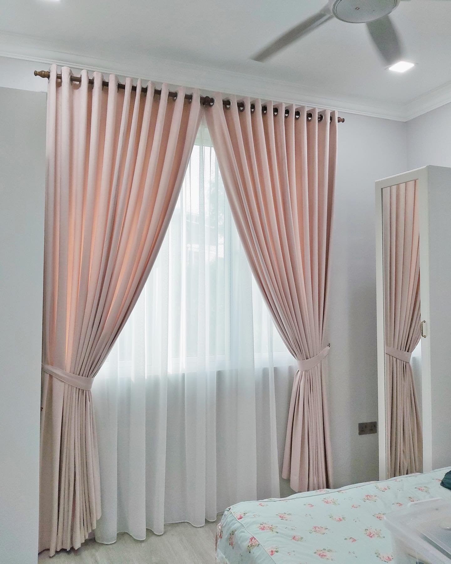 Blush pink grommet curtains tied back over white sheer curtains in a bright bedroom.