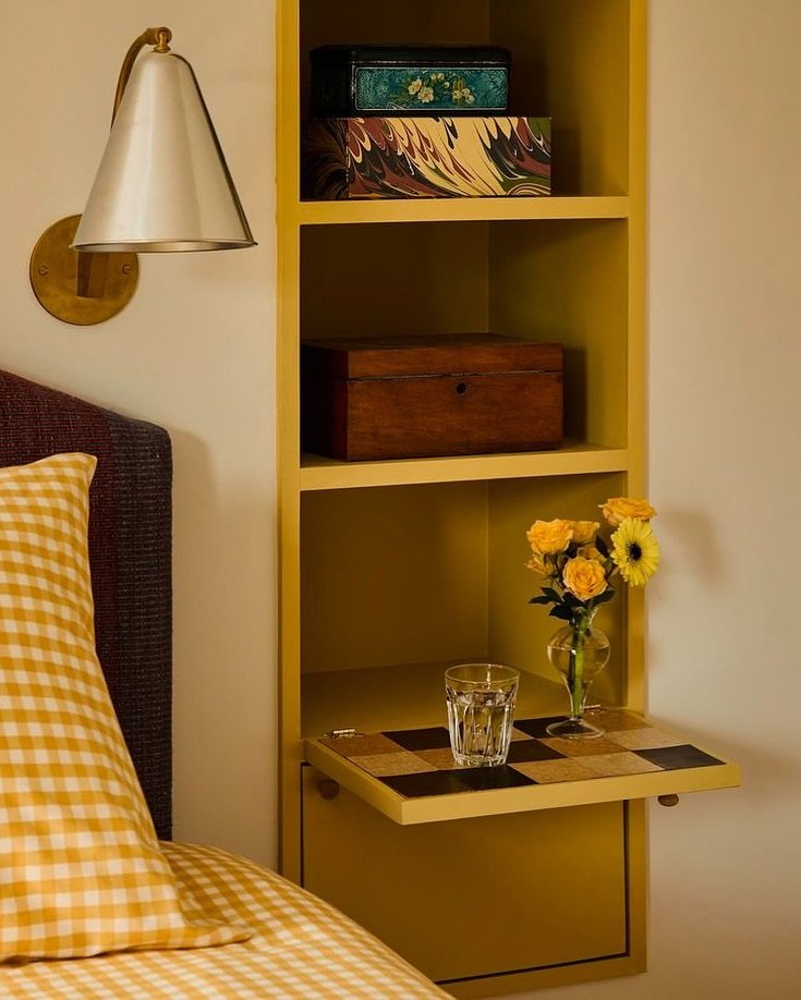 A narrow, floor-to-ceiling yellow storage tower next to a bed, with open shelving and a checkerboard drop-down shelf used as a small nightstand.