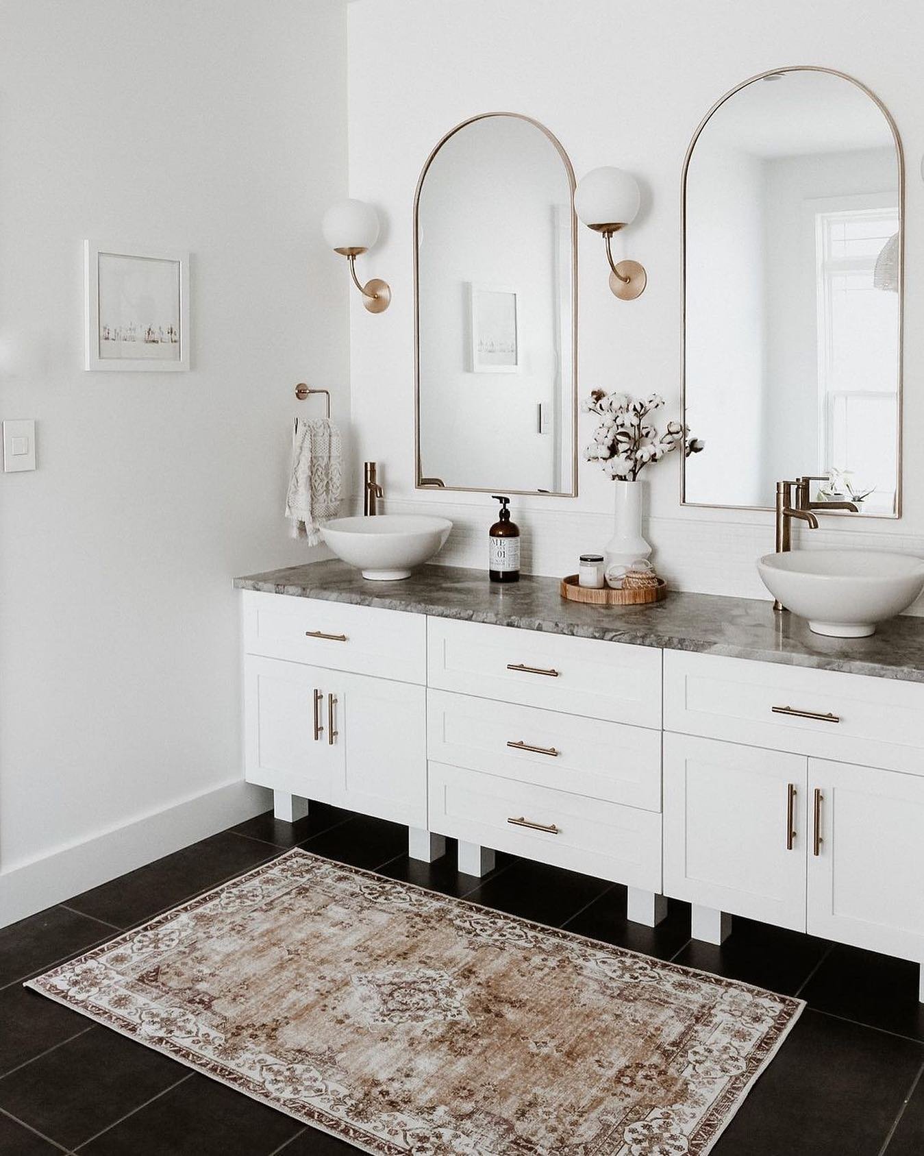 A bright white bathroom with a long double vanity, dark gray tile floor, arched gold-framed mirrors, and a rectangular distressed beige and brown patterned rug.