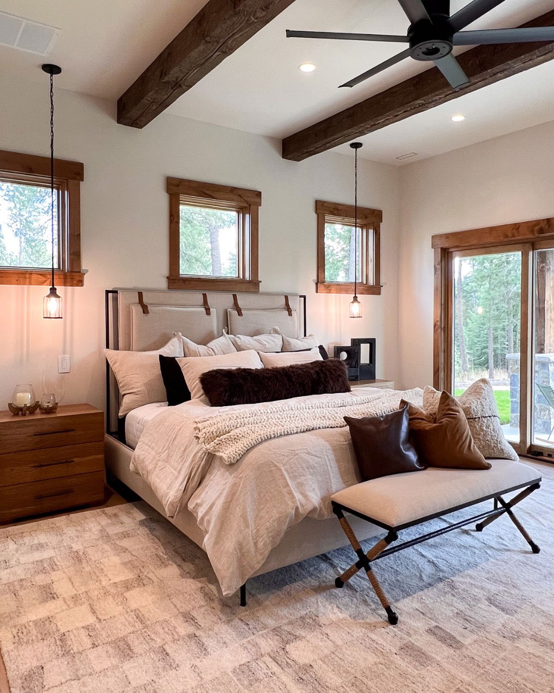 A cozy bedroom featuring exposed wood ceiling beams, a bed with neutral linens, and a long upholstered bench with dark metal X-shaped legs placed at the foot of the bed.
